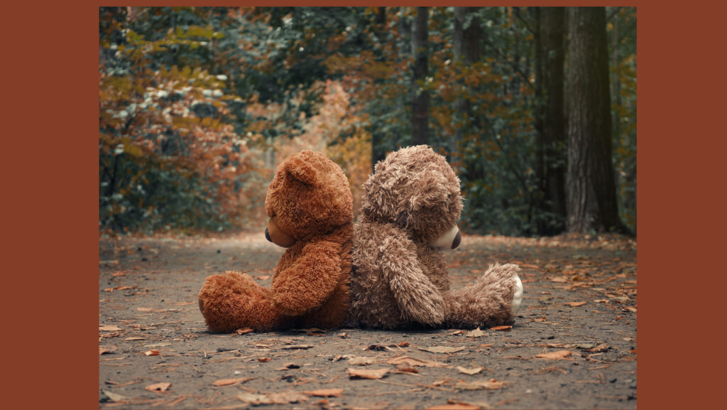 two teddy bears sitting back to back in the middle of an autumn forest.
